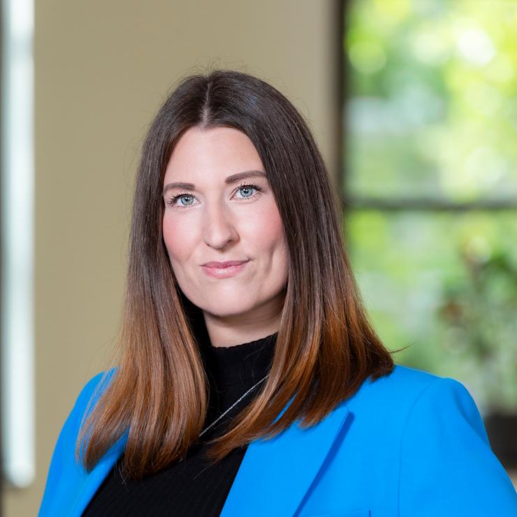 Frau mit langen braunen Haaren und blauem Blazer steht vor einem Fenster mit Blick ins Grüne.