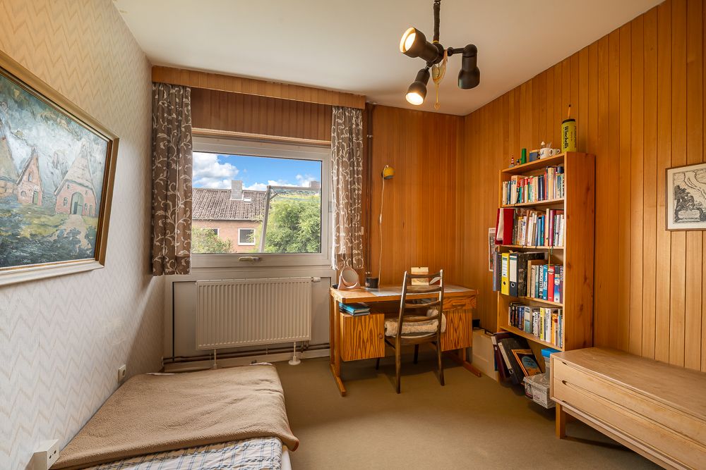 Kleines Zimmer mit Schreibtisch, Bücherregal, Bett und Fenster, Holzwände und heller Teppichboden.