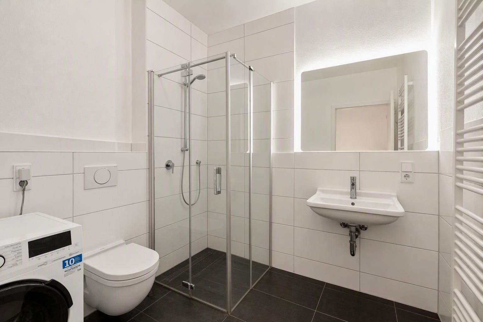 Modernes Bad mit Dusche, Waschbecken, Wandtoilette, Waschmaschine und beleuchtetem Spiegel.