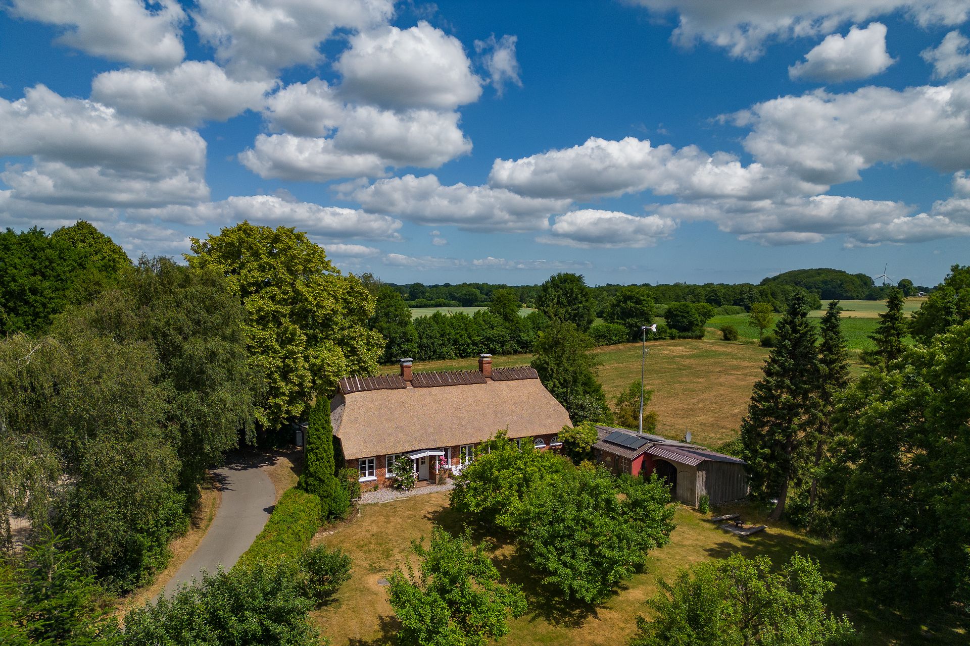 Reetgedecktes Haus mit großem Garten, umgeben von Bäumen und Feldern unter blauem Himmel.