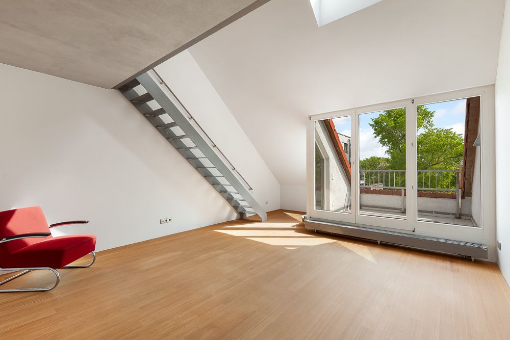 Helles Zimmer mit Holzfußboden, rotem Sessel, Metalltreppe und großer Fensterfront zur Terrasse.