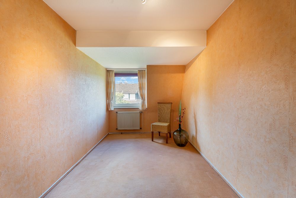 Schmaler Raum mit beigem Teppich, Stuhl am Fenster und großer Vase mit Zweigen.