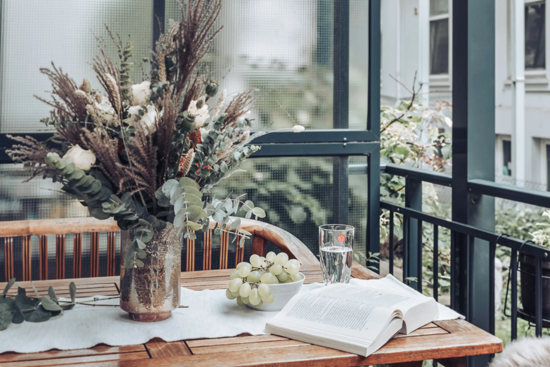 Holztisch auf Balkon mit Blumenstrauß, Glas Wasser, Weintrauben und offenem Buch.