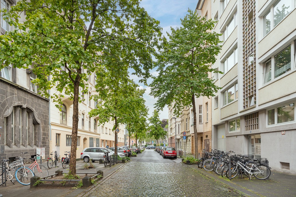 Baumgesäumte Wohnstraße mit parkenden Autos und vielen Fahrrädern vor Wohnhäusern.