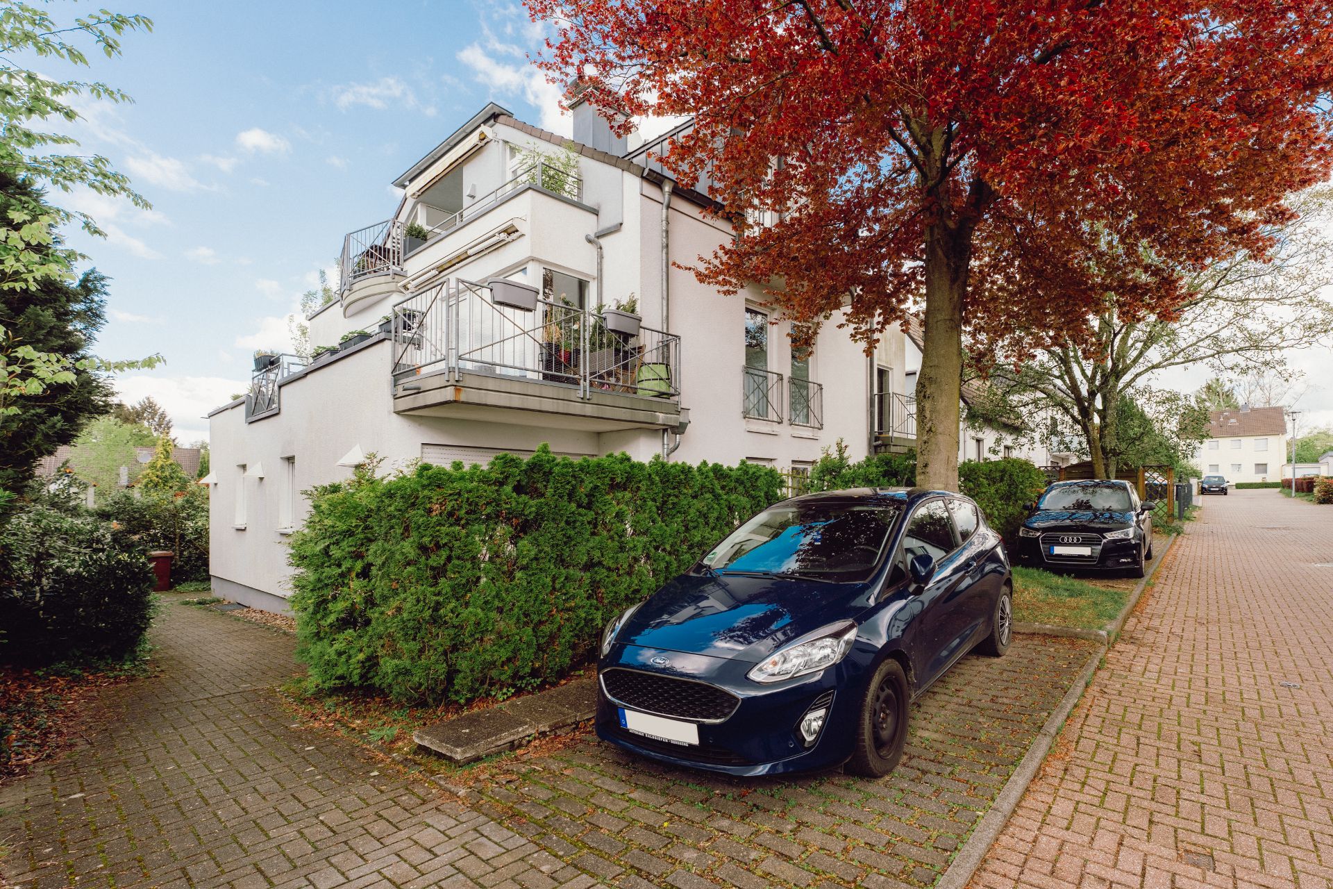 Modernes Wohnhaus mit Balkonen, davor parken zwei Autos am Straßenrand unter Bäumen.