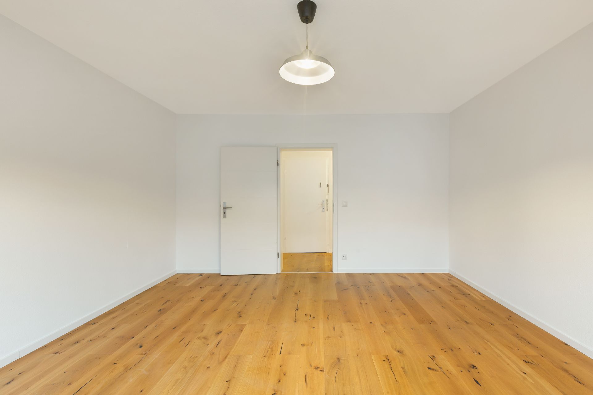 Leerer Raum mit Holzboden, weißer Wand, Tür und Deckenlampe.