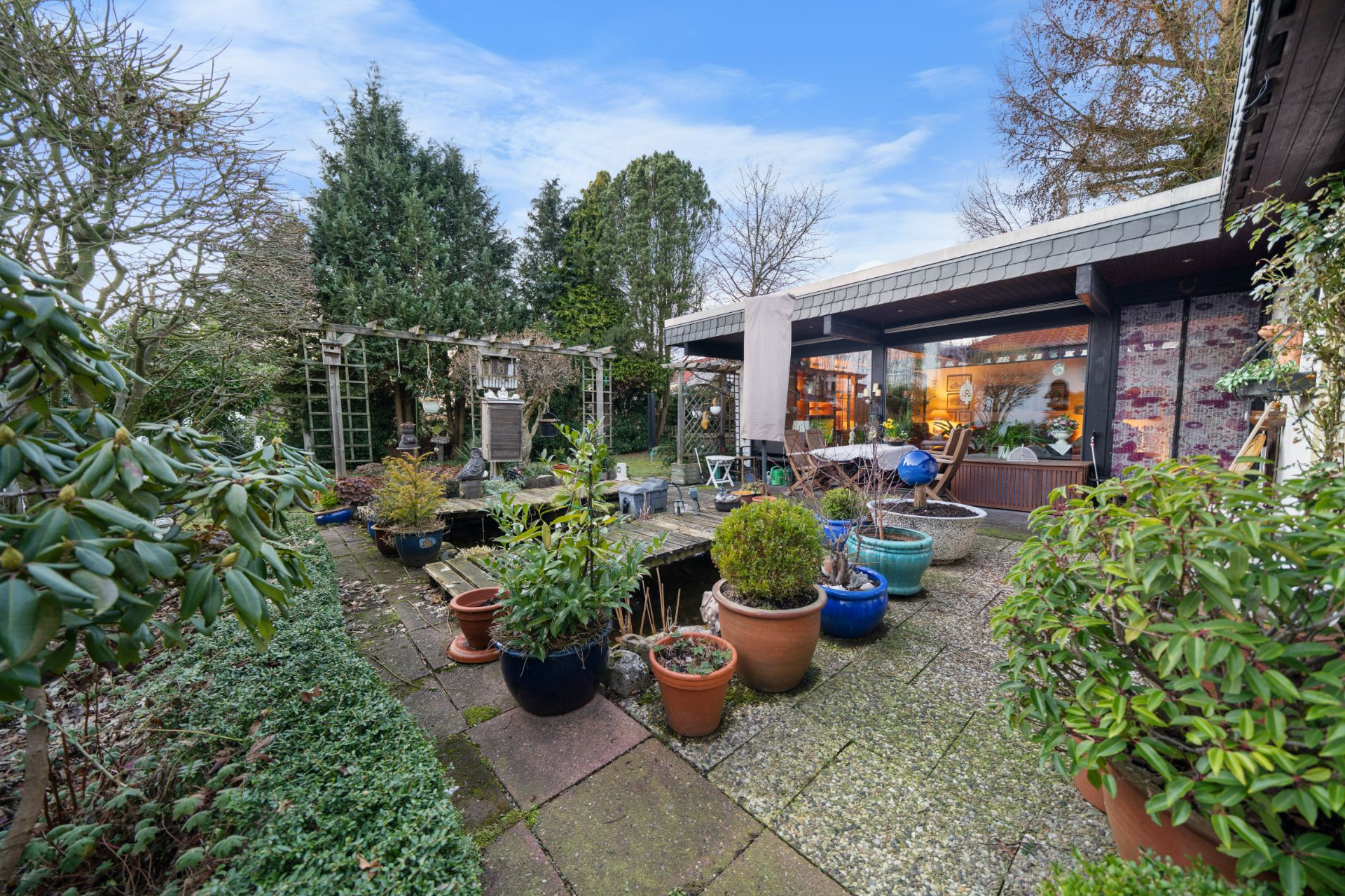 Gepflasterte Terrasse mit vielen Blumentöpfen, Pflanzen und Blick auf ein Wohnhaus mit großen Fenstern.