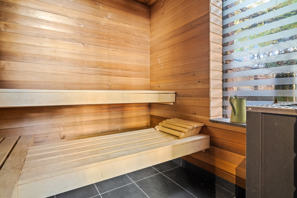 Holzsauna mit zwei Bänken, einem Fenster mit Lamellen und einem grünen Eimer auf der Fensterbank.