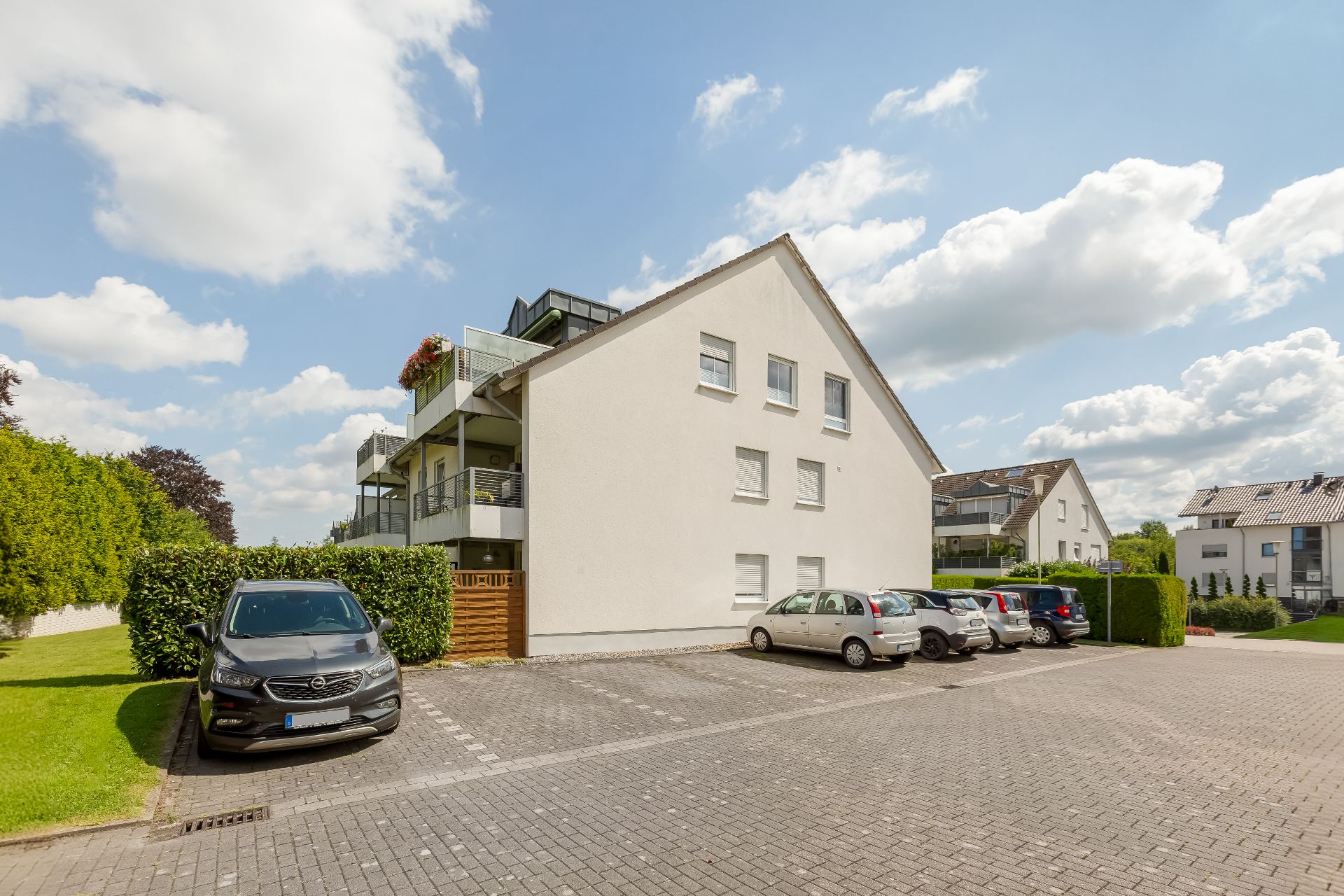 Mehrfamilienhaus mit Balkonen und Parkplatz, mehrere Autos stehen auf gepflastertem Hof.