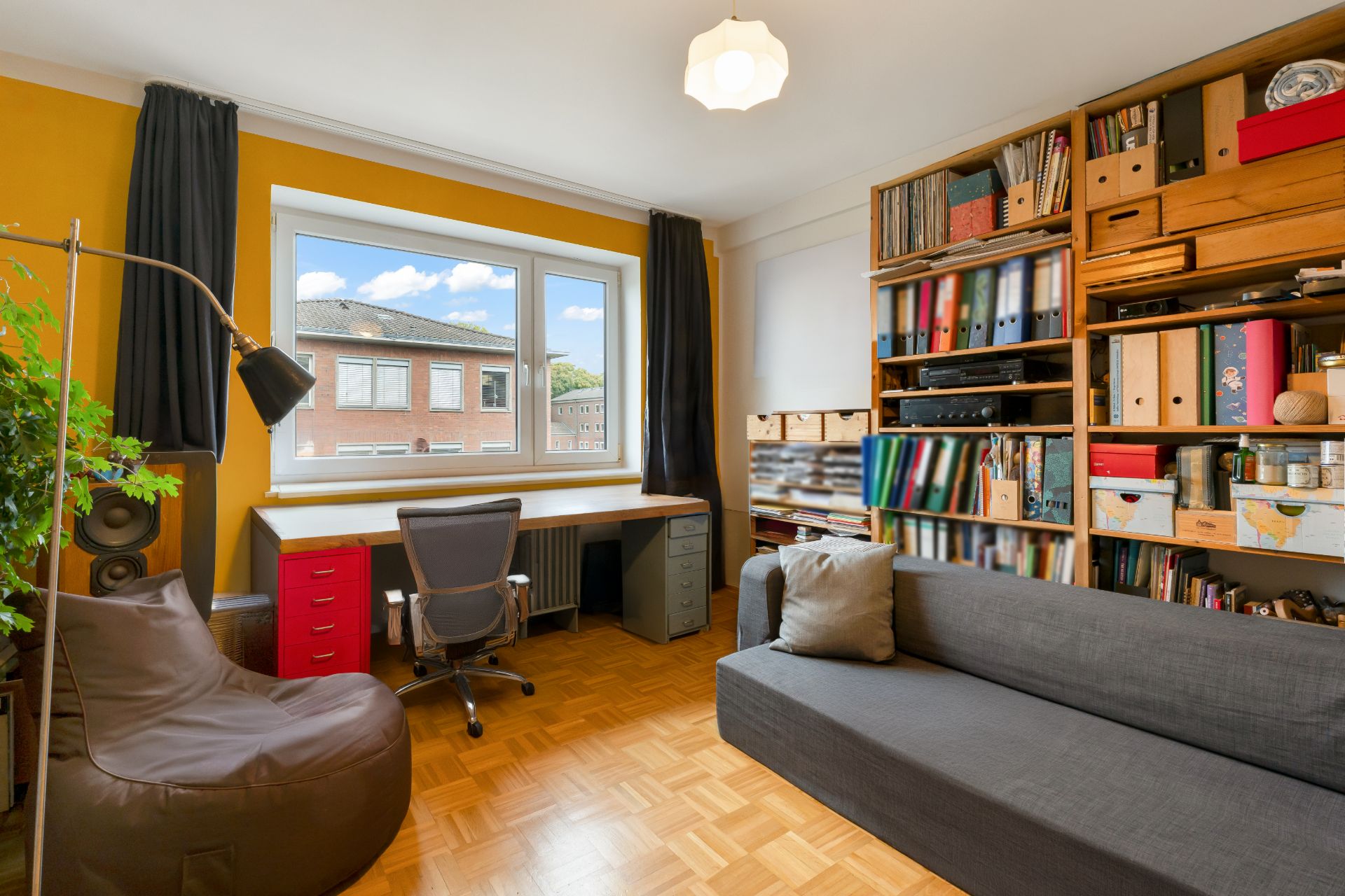 Hell eingerichtetes Arbeitszimmer mit Schreibtisch, Bürostuhl, Sofa und Regalen voller Ordner und Bücher.