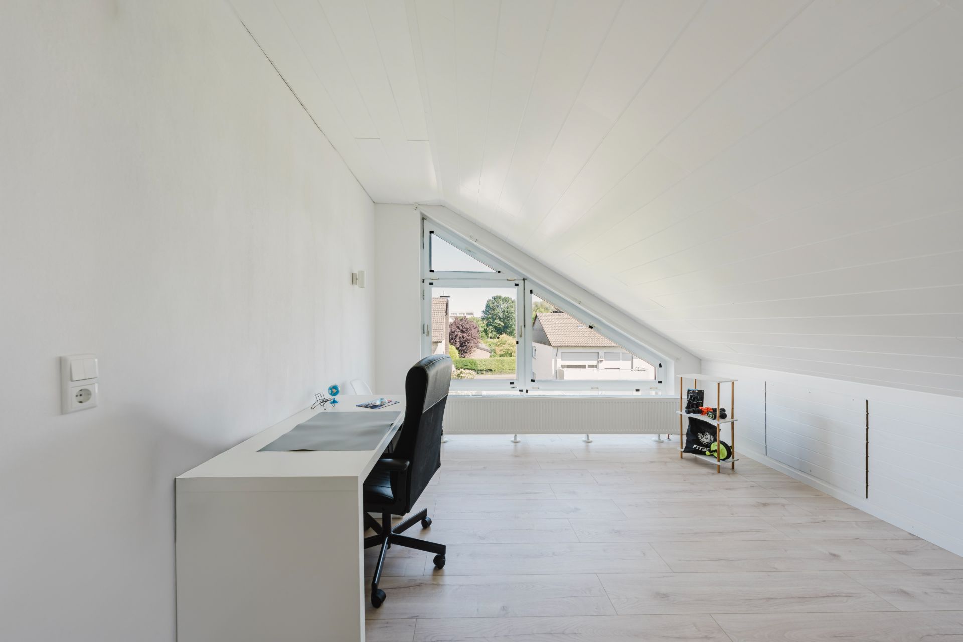 Helles Dachzimmer mit Schreibtisch, Stuhl und großem Fenster zur Straße.