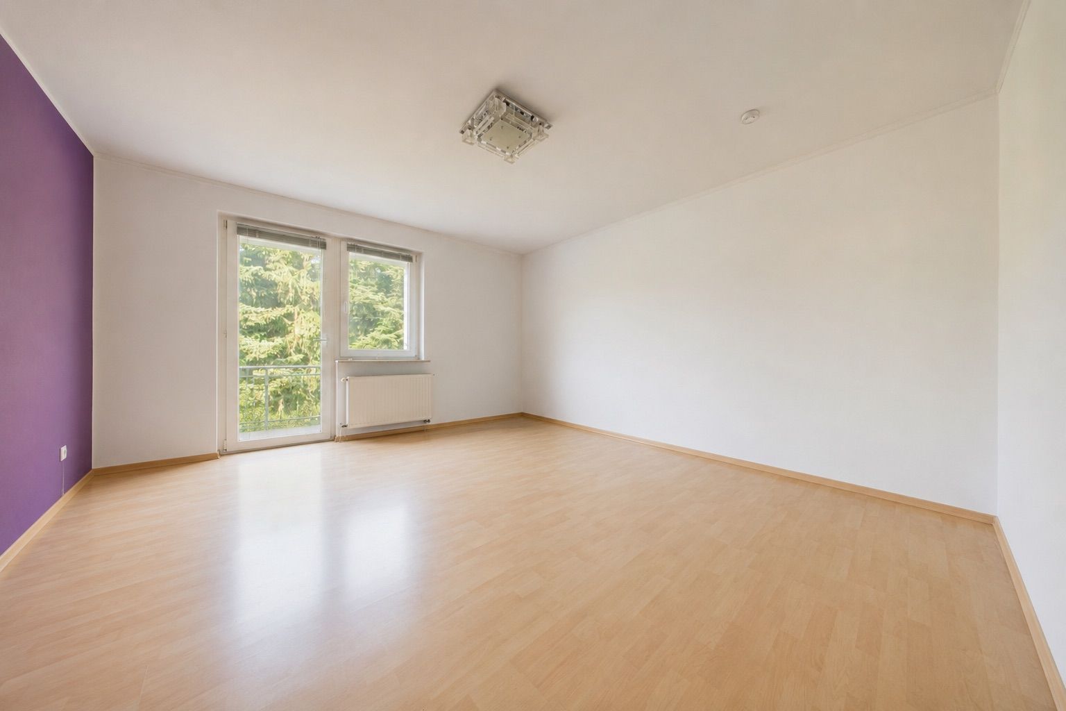 Leeres Zimmer mit hellem Holzboden, weißen Wänden und einem lila Akzent, Fenster mit Blick aufs Grün.