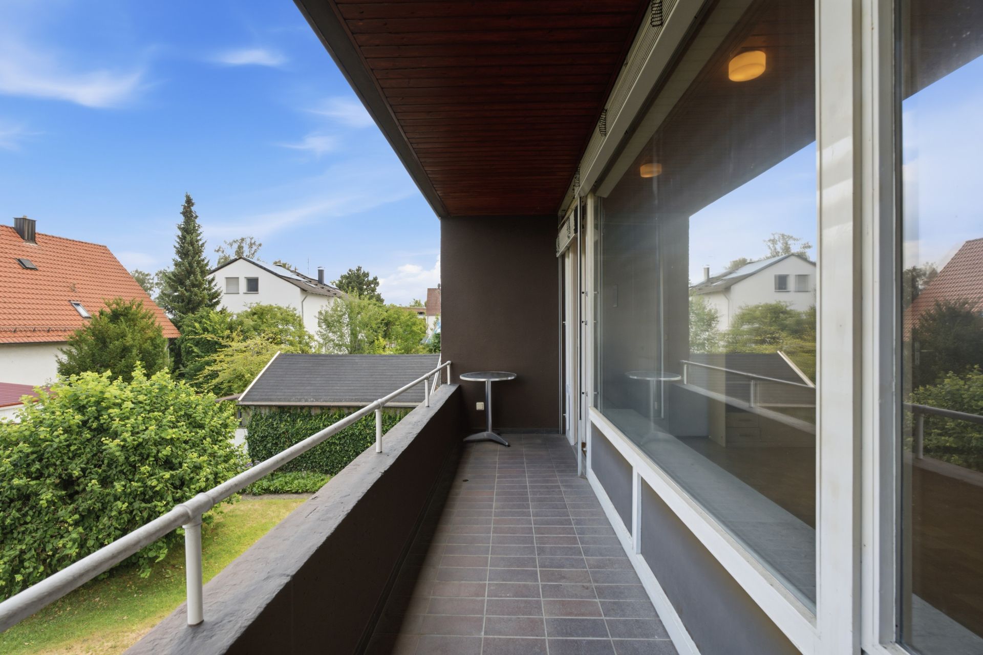Langer Balkon mit Fliesenboden, Geländer und kleinem Tisch, Blick auf Nachbarhäuser und Gärten.