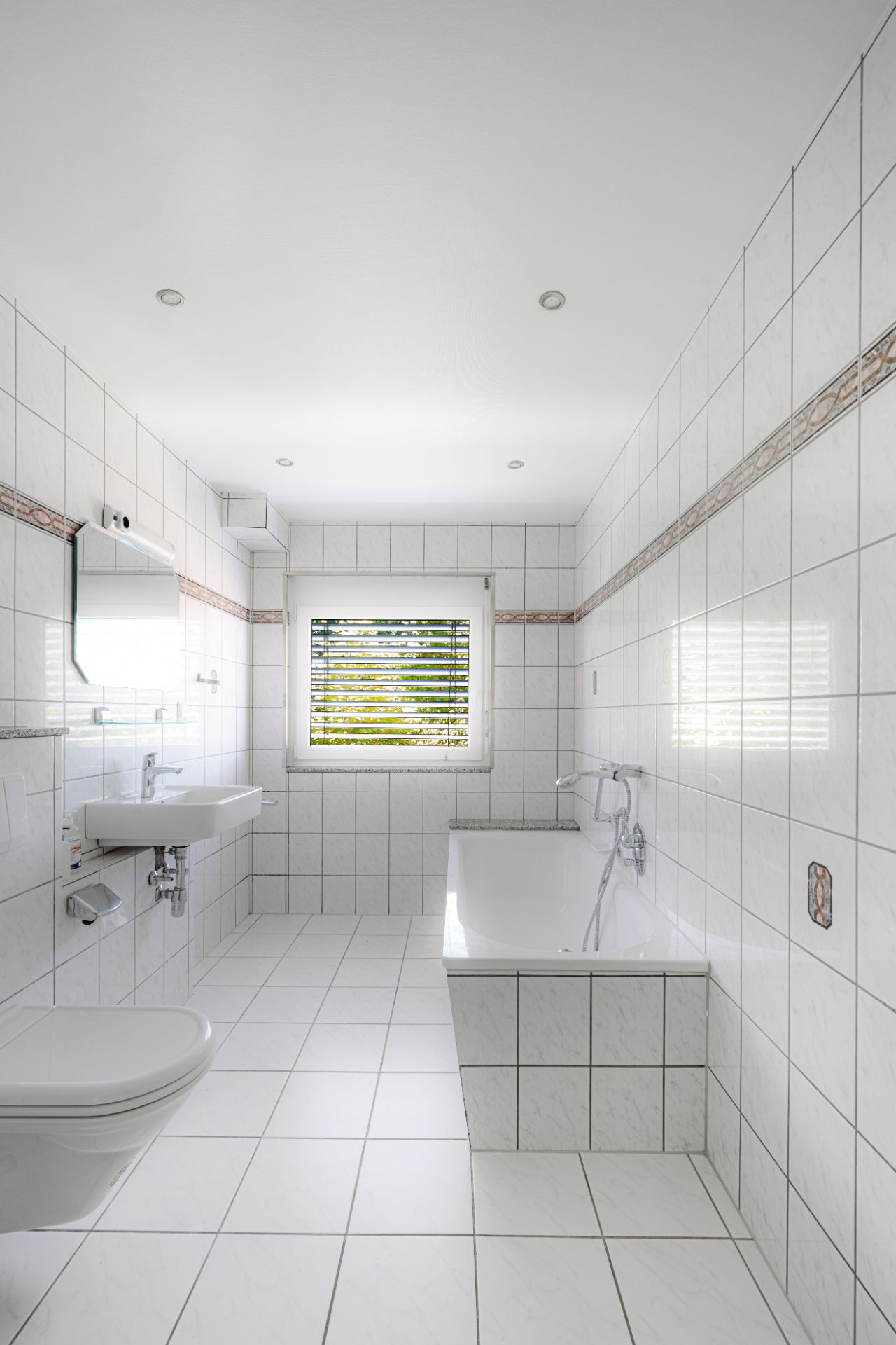 Helles Badezimmer mit Badewanne, Waschbecken, Toilette und Fenster, alles gefliest.