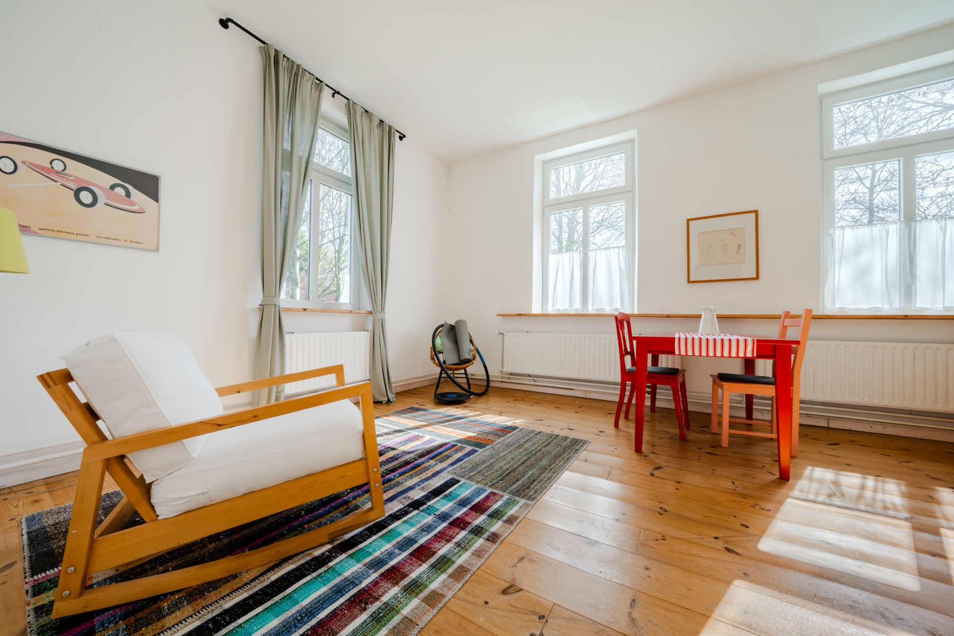 Helles Wohnzimmer mit Sessel, kleinem Tisch, Stühlen und buntem Teppich auf Holzboden.
