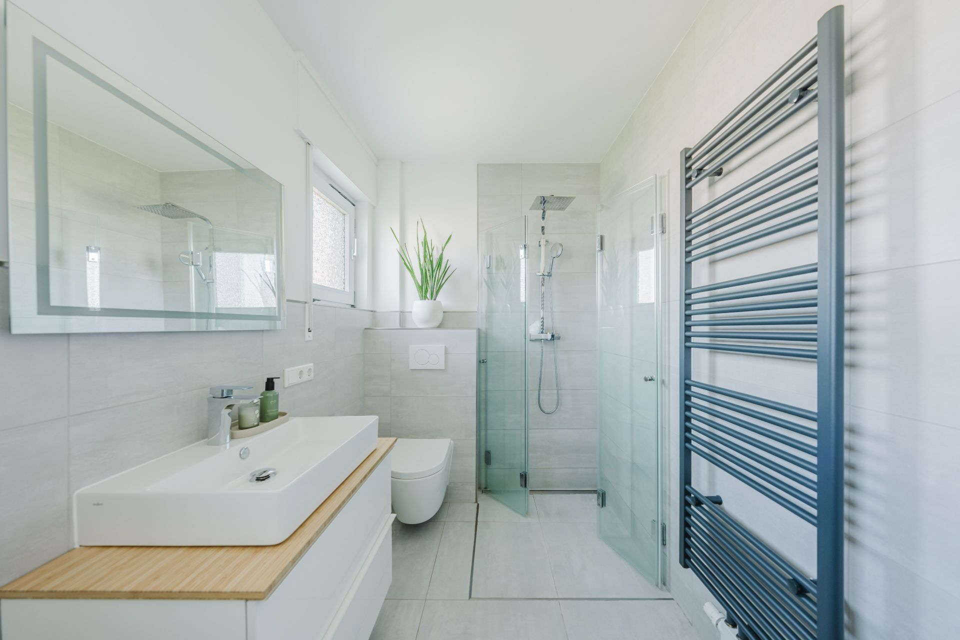 Modernes Bad mit großer Dusche, Waschbecken, Spiegel, WC, Handtuchheizkörper und Pflanze am Fenster.