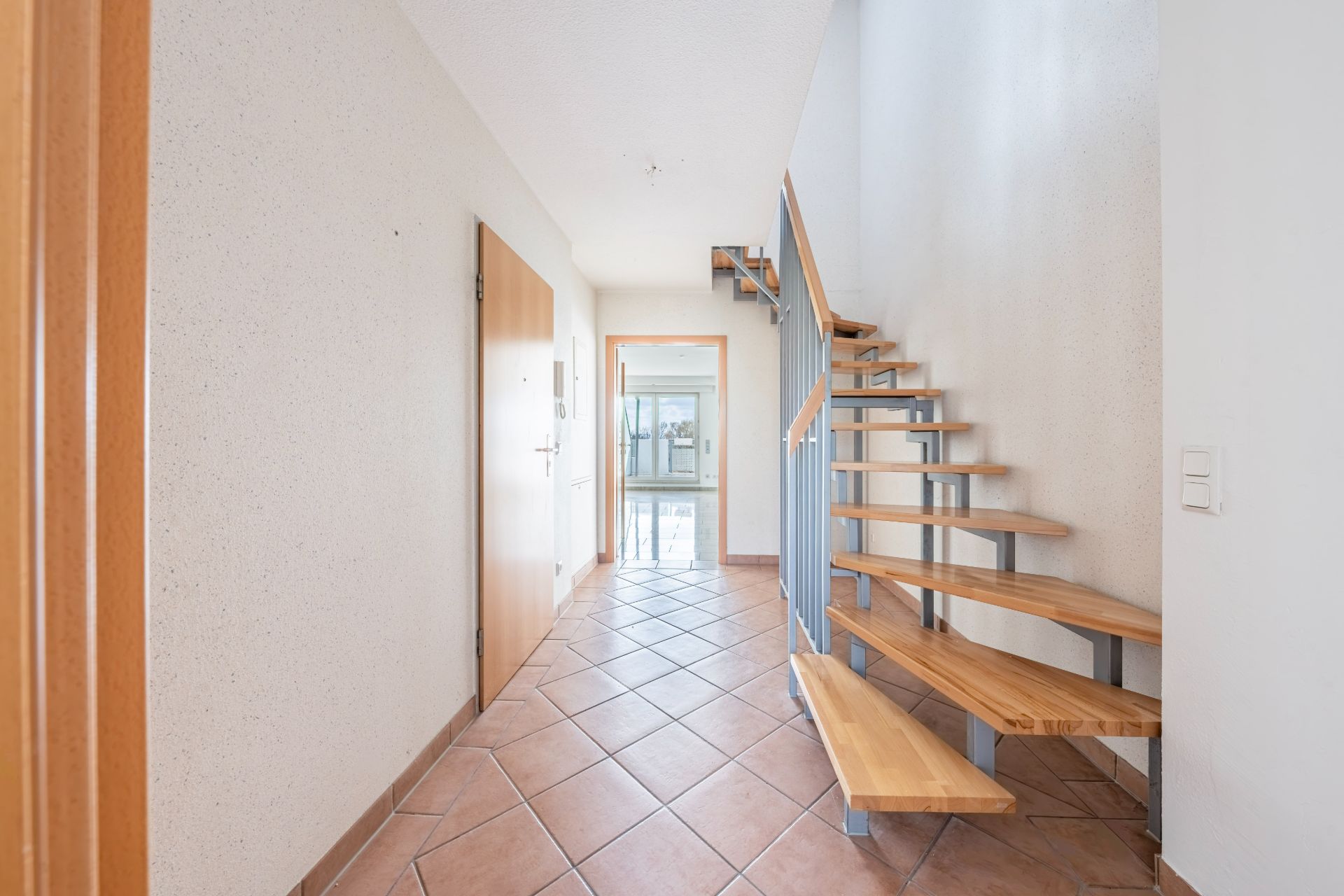 Heller Flur mit gefliesten Boden und moderner Holztreppe, Tür am Ende des Flurs.