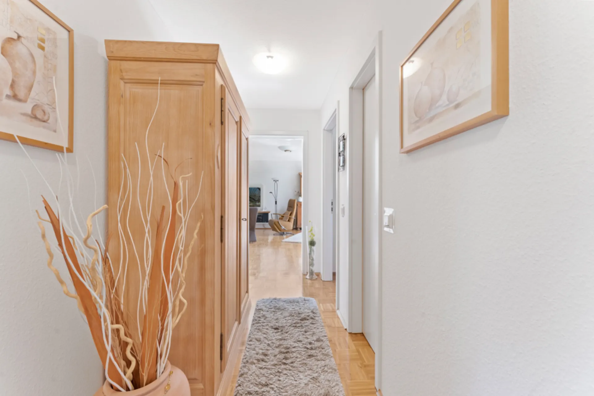 Heller Flur mit Teppich, Holzschrank, Trockenpflanzen und Bildern an den Wänden.