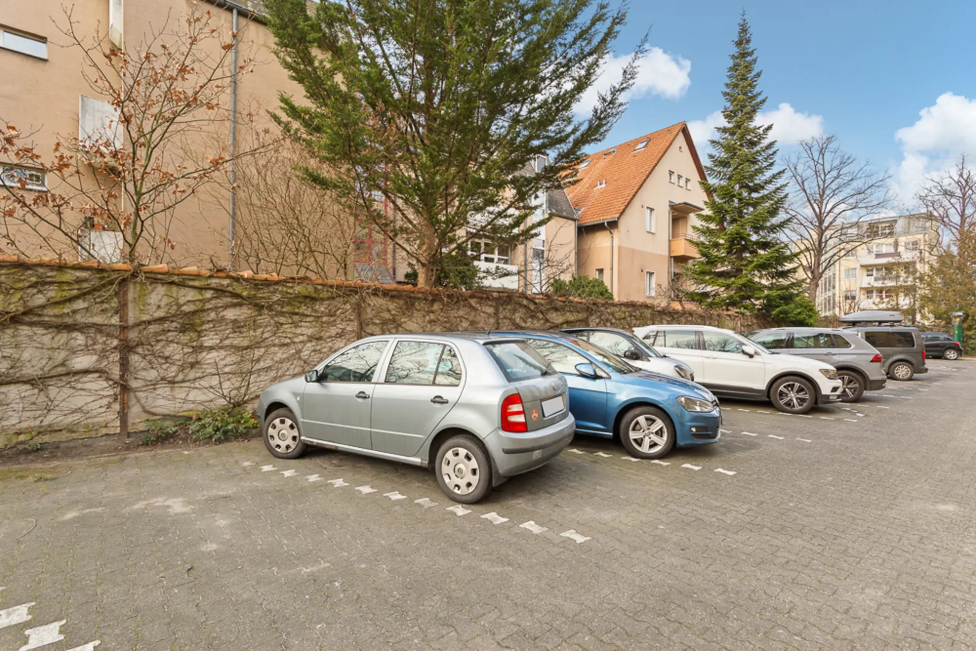 Parkplatz mit mehreren Autos neben Wohngebäuden, Bäume und Mauer im Hintergrund.