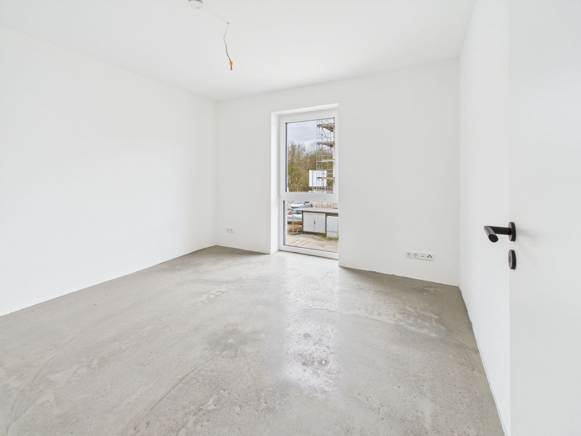 Leeres Zimmer mit weiß gestrichenen Wänden, Betonboden und einem Fenster mit Tür nach außen.