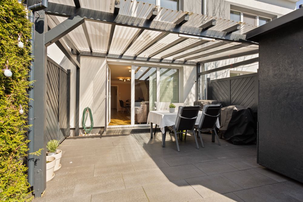 Überdachte Terrasse mit Esstisch, Stühlen und Grill neben einem Wohnhaus.
