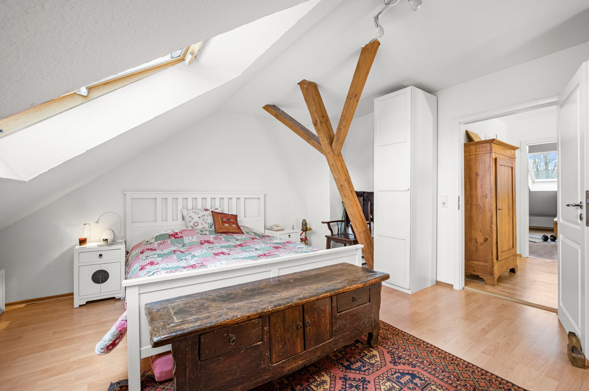 Helles Schlafzimmer mit Dachfenster, Doppelbett, Holztruhe und sichtbarem Holzbalken.