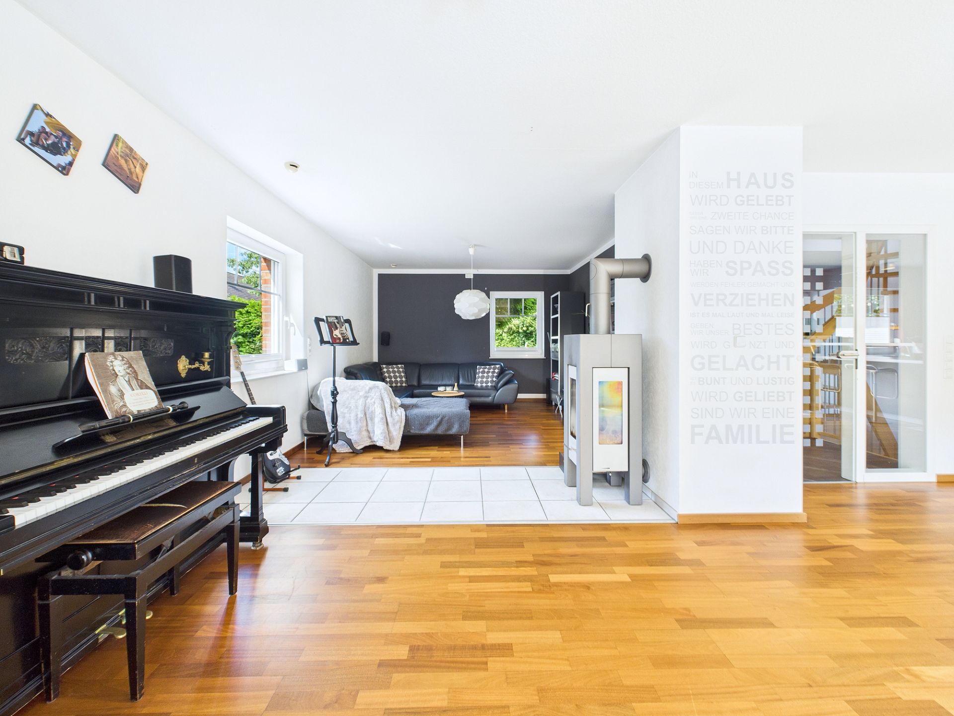 Helles Wohnzimmer mit Klavier, Sofa, Ofen und großem Fenster auf Parkettboden.