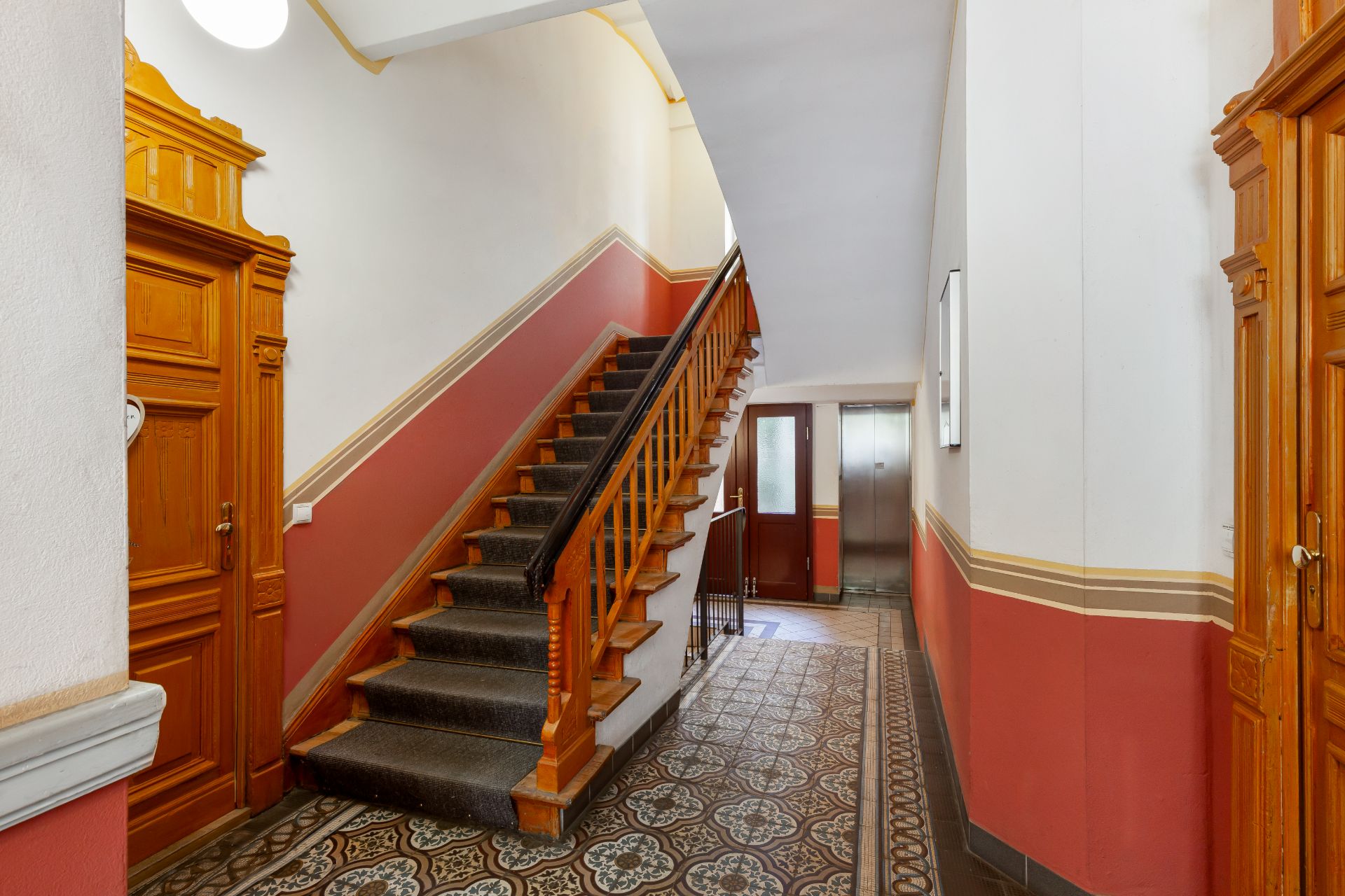 Hausflur mit gemustertem Fliesenboden, Holztreppe und Aufzug im Hintergrund.