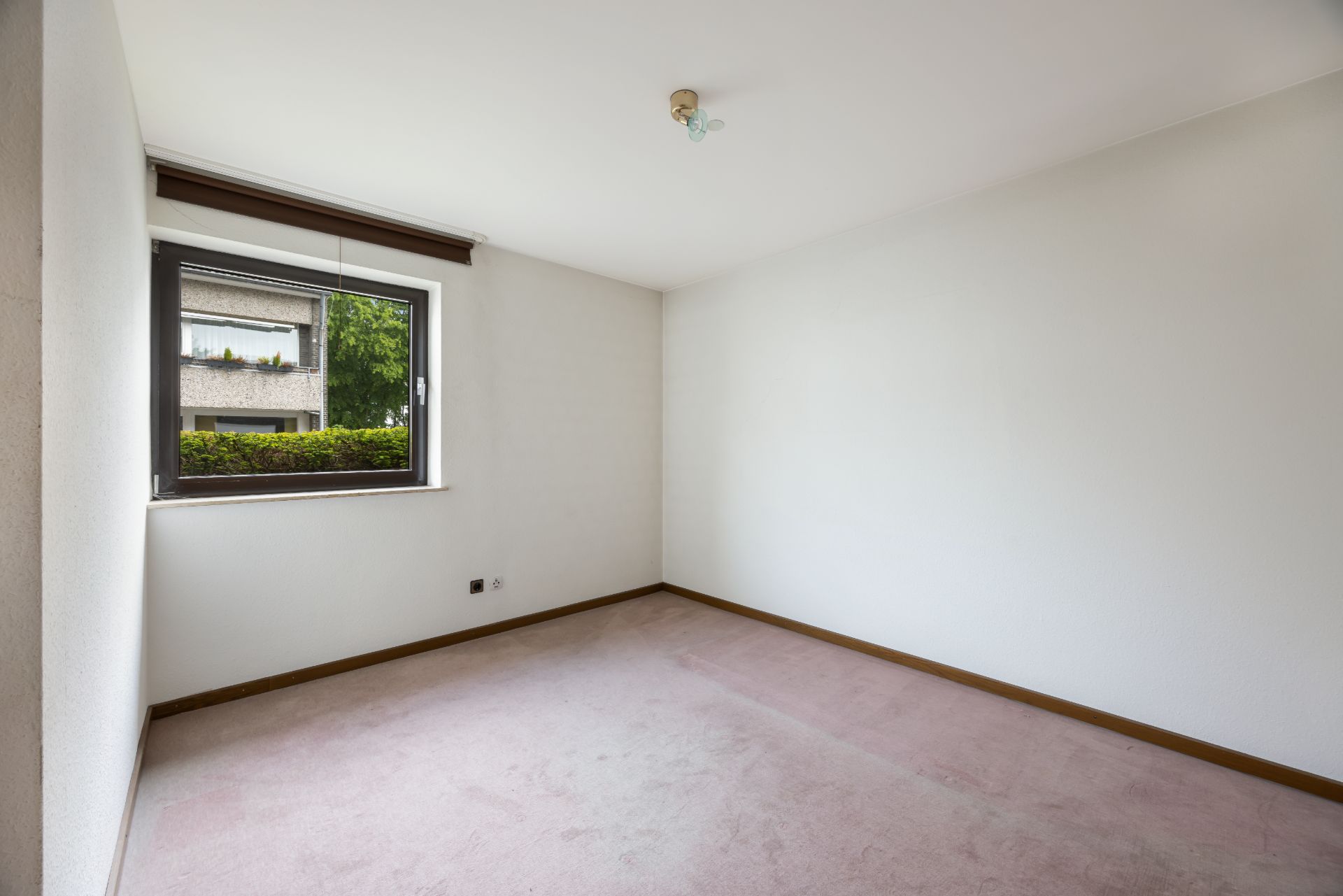 Leeres Zimmer mit Teppichboden, weiß gestrichenen Wänden und einem Fenster mit Blick nach draußen.