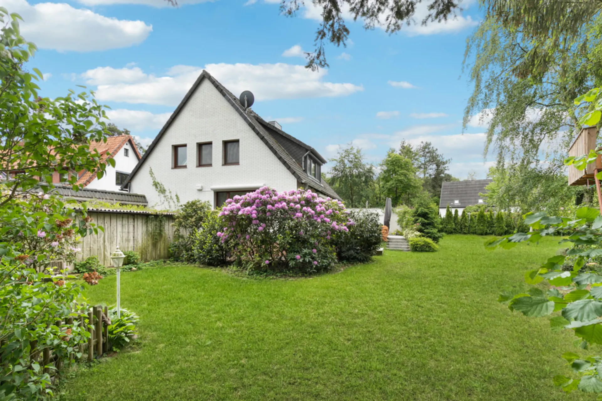 Haus mit Satteldach und großem Garten mit Blumen und Rasen unter blauem Himmel.
