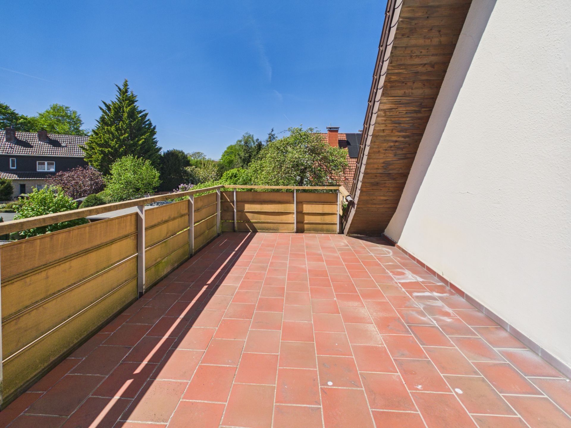 Große Terrasse mit roten Fliesen, Holzgeländer und Blick auf Bäume und Nachbarhäuser.
