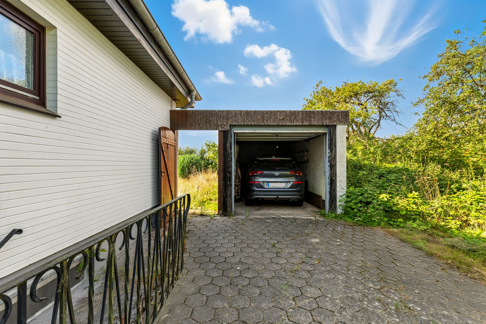 Auto steht in einer offenen Garage neben einem Wohnhaus, umgeben von Garten und Büschen.