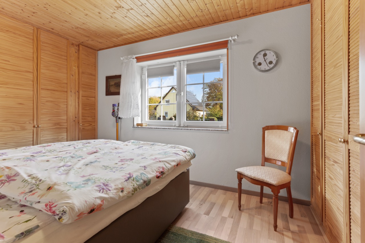 Helles Schlafzimmer mit Doppelbett, Holzschränken, Stuhl und Fenster mit Ausblick.