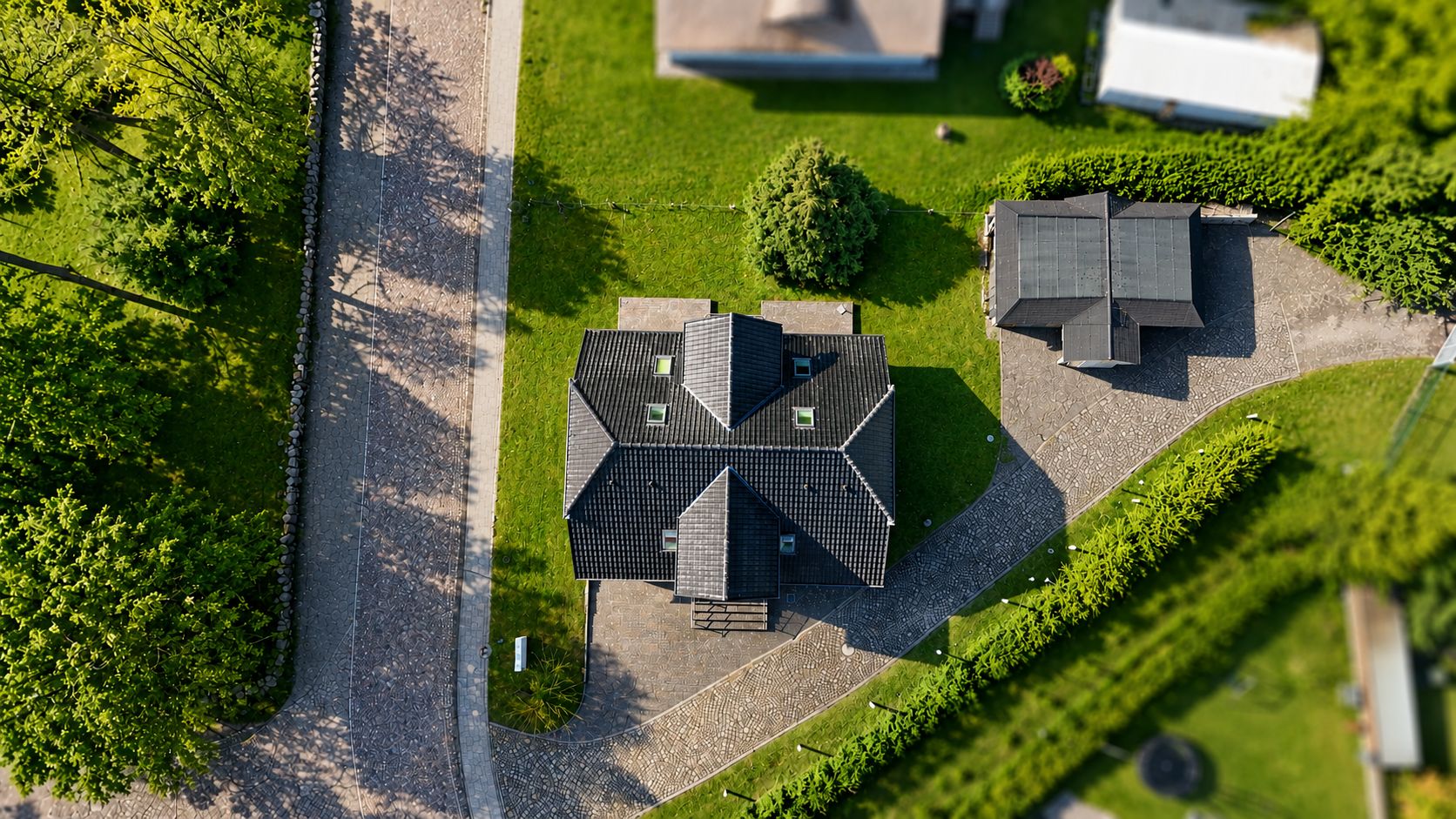 Luftaufnahme eines Hauses mit dunklem Dach, Garten und Nebenhaus an einer gepflasterten Straße.