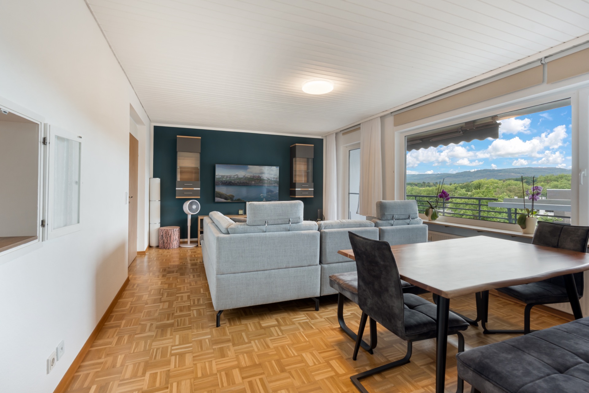 Helles Wohnzimmer mit Essbereich, grauen Sofas, großem Fenster und Holzboden.