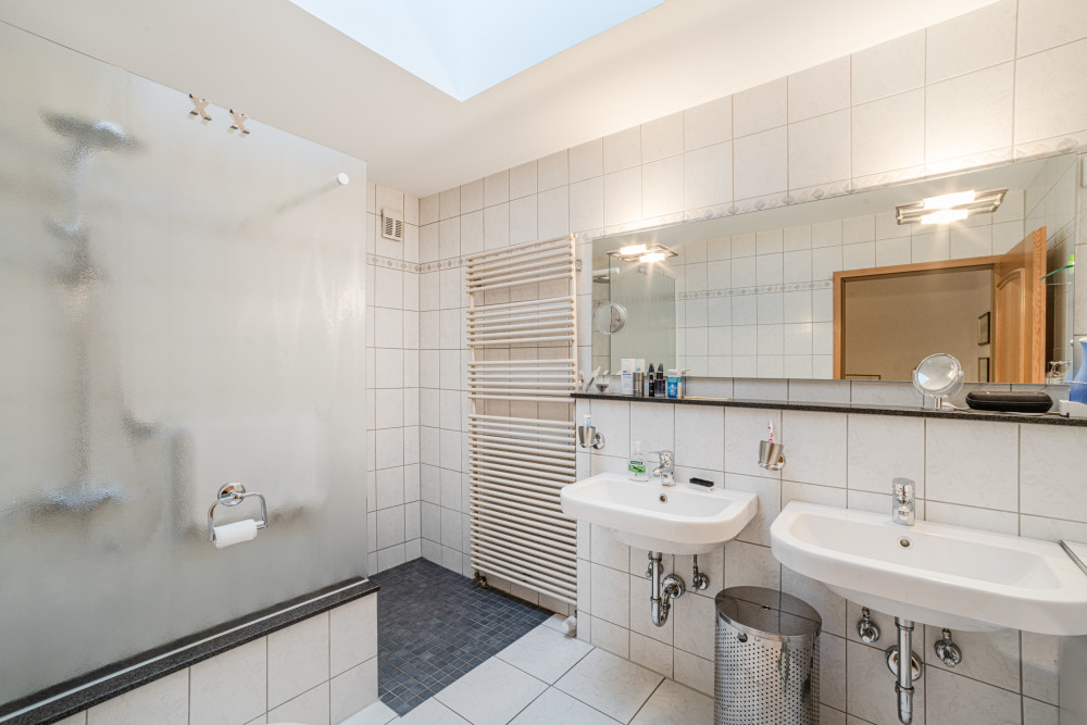 Helles Badezimmer mit zwei Waschbecken, großem Spiegel, Dusche und Handtuchheizung.