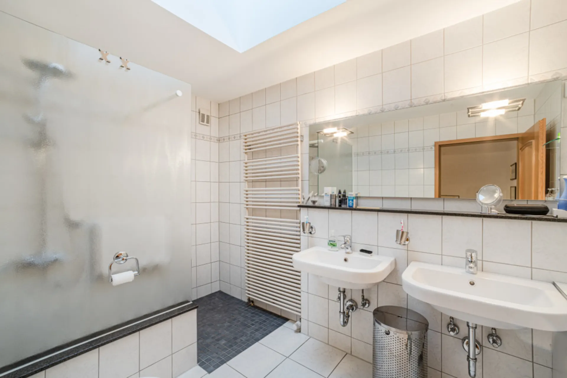 Helles Badezimmer mit zwei Waschbecken, großem Spiegel, Dusche und Handtuchheizung.