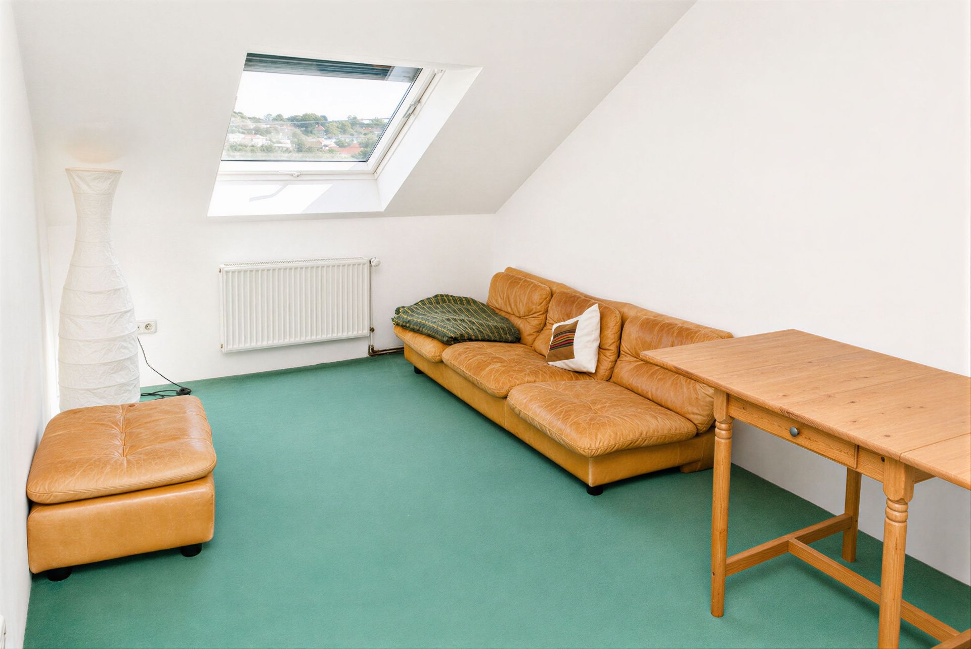 Dachzimmer mit grünem Teppich, braunem Sofa, Ottomane, Stehlampe, Tisch und Dachfenster.