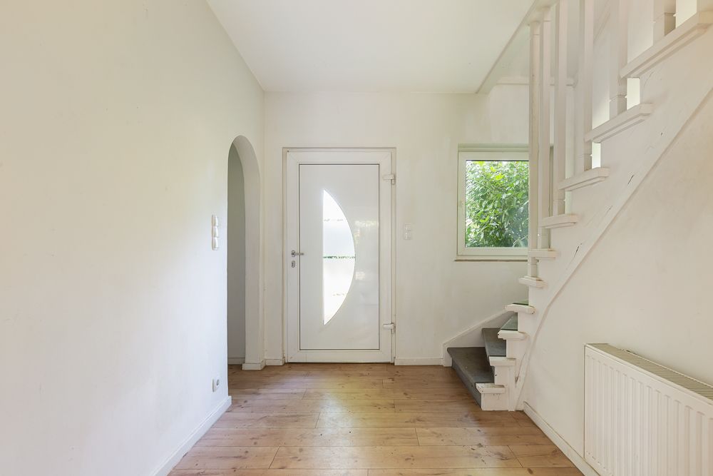 Heller Flur mit Holzboden, weißer Haustür, Fenster und Treppe nach oben.