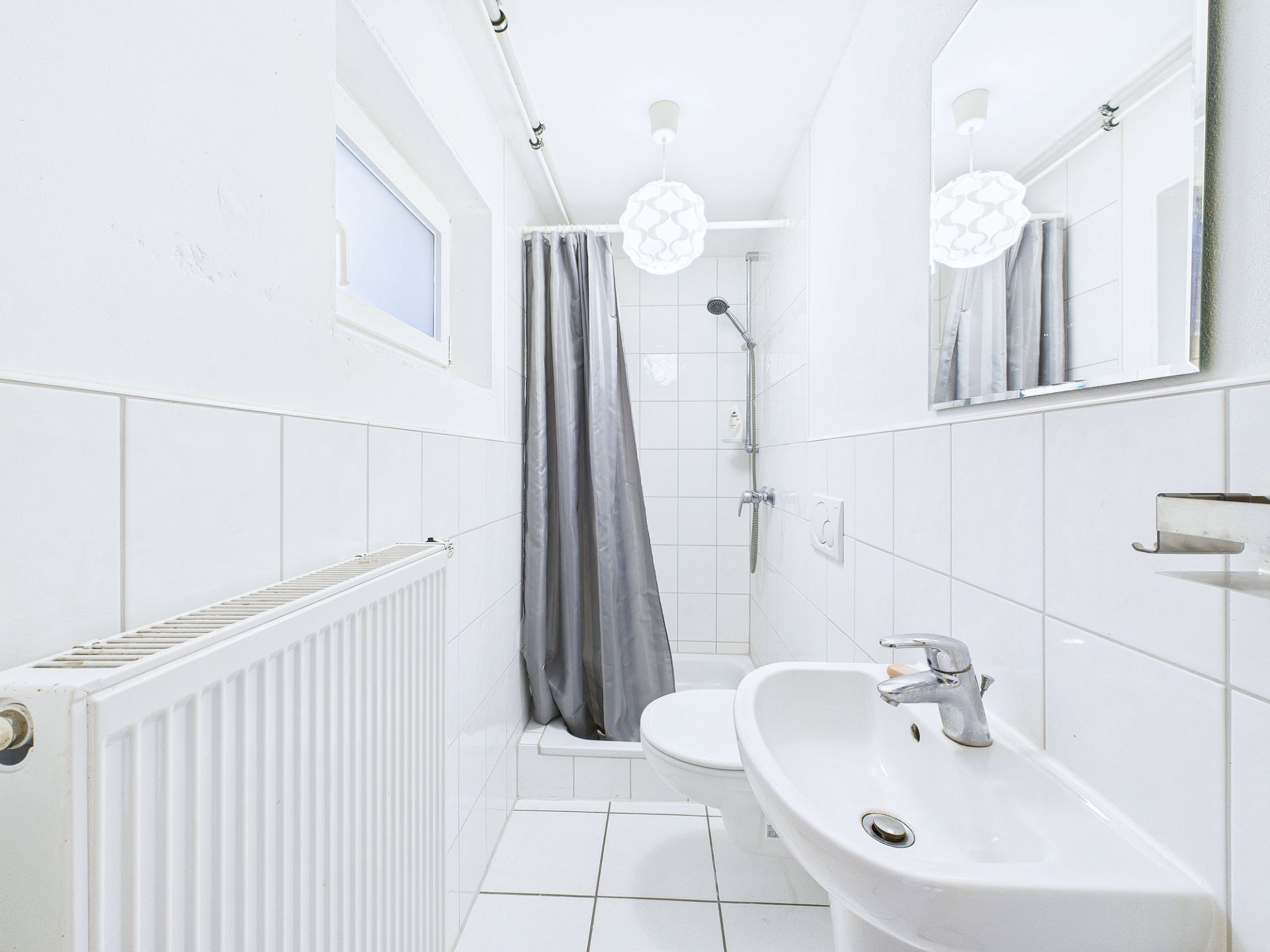 Helles, schmales Badezimmer mit Dusche, weißem Waschbecken, Spiegel und grauem Duschvorhang.