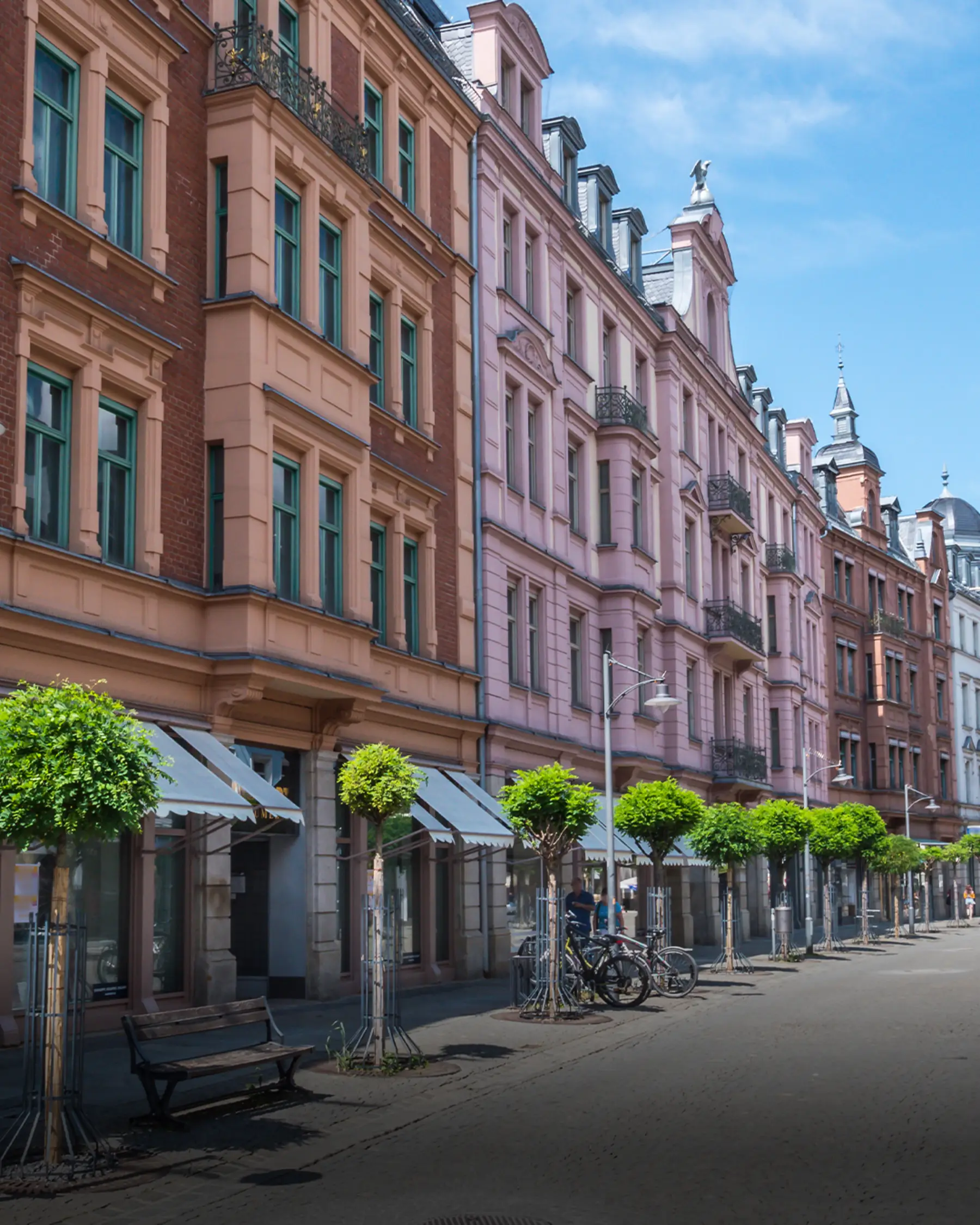 Mehrstöckige Wohnhäuser mit grünen Fenstern, Bäumen und Fahrrädern an einer ruhigen Straße.