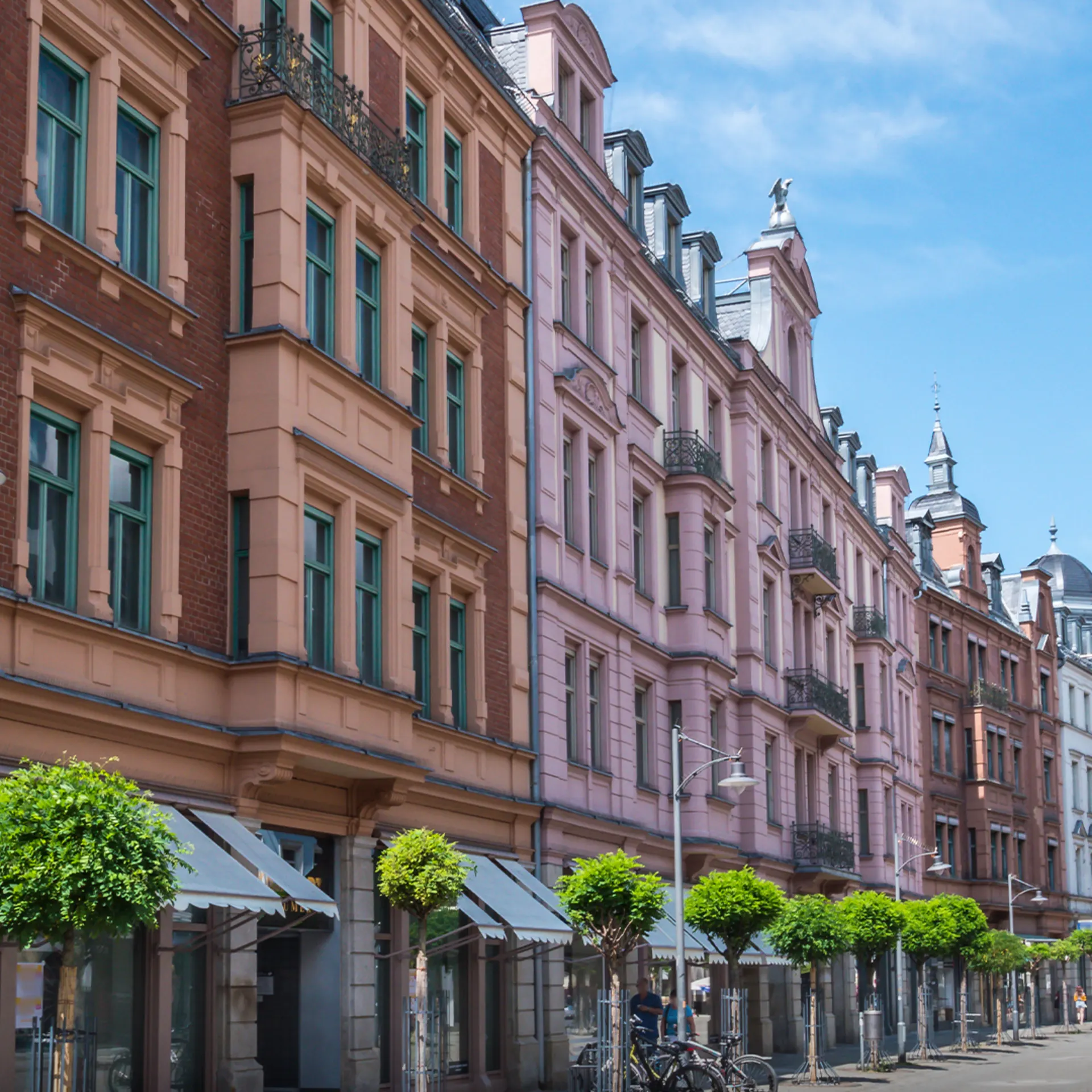 Mehrstöckige Wohnhäuser mit grünen Fenstern, Bäumen und Fahrrädern an einer ruhigen Straße.