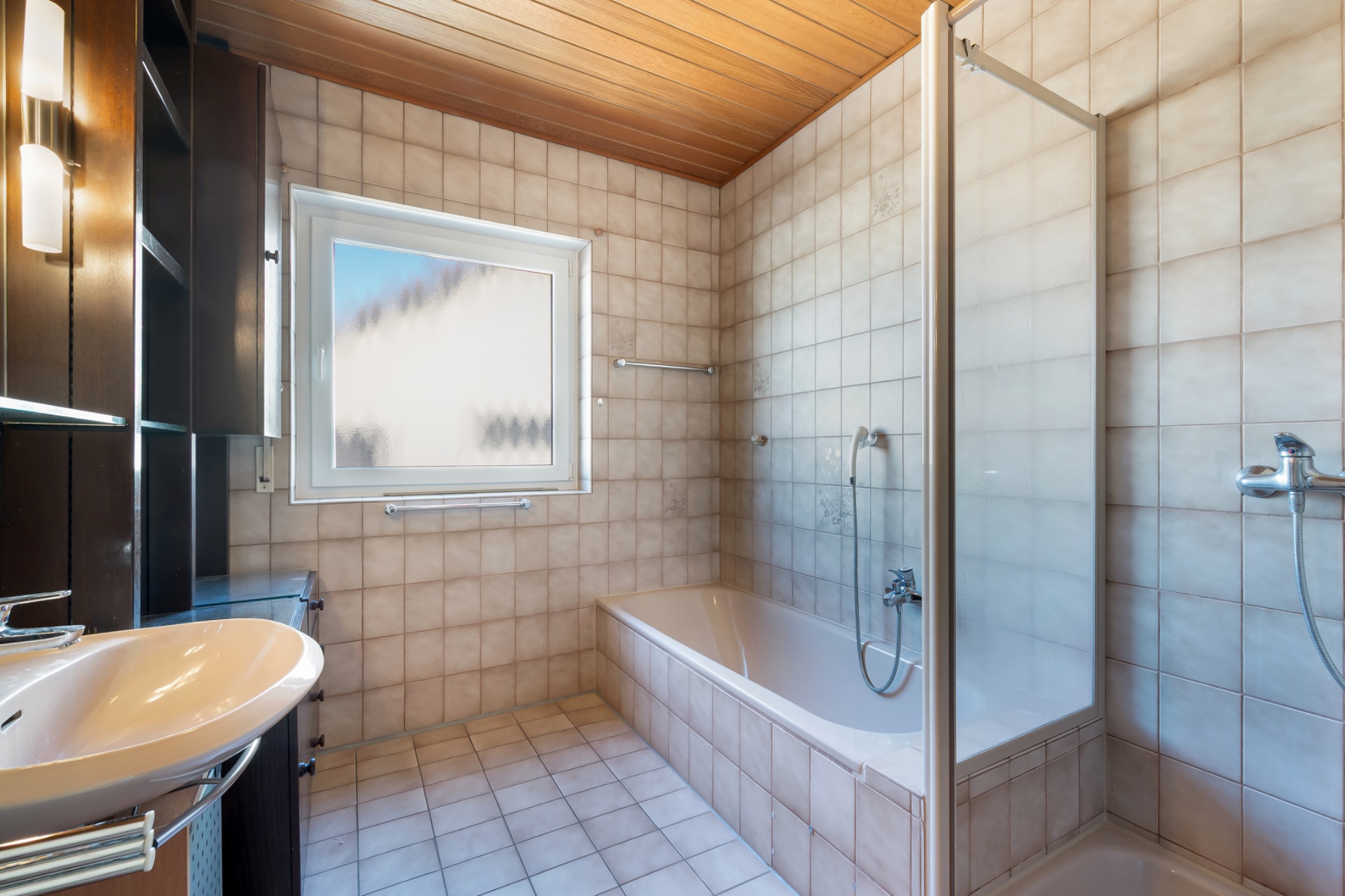 Helles Badezimmer mit Badewanne, Dusche, Waschbecken und Fenster, gefliest und mit Holzdecke.