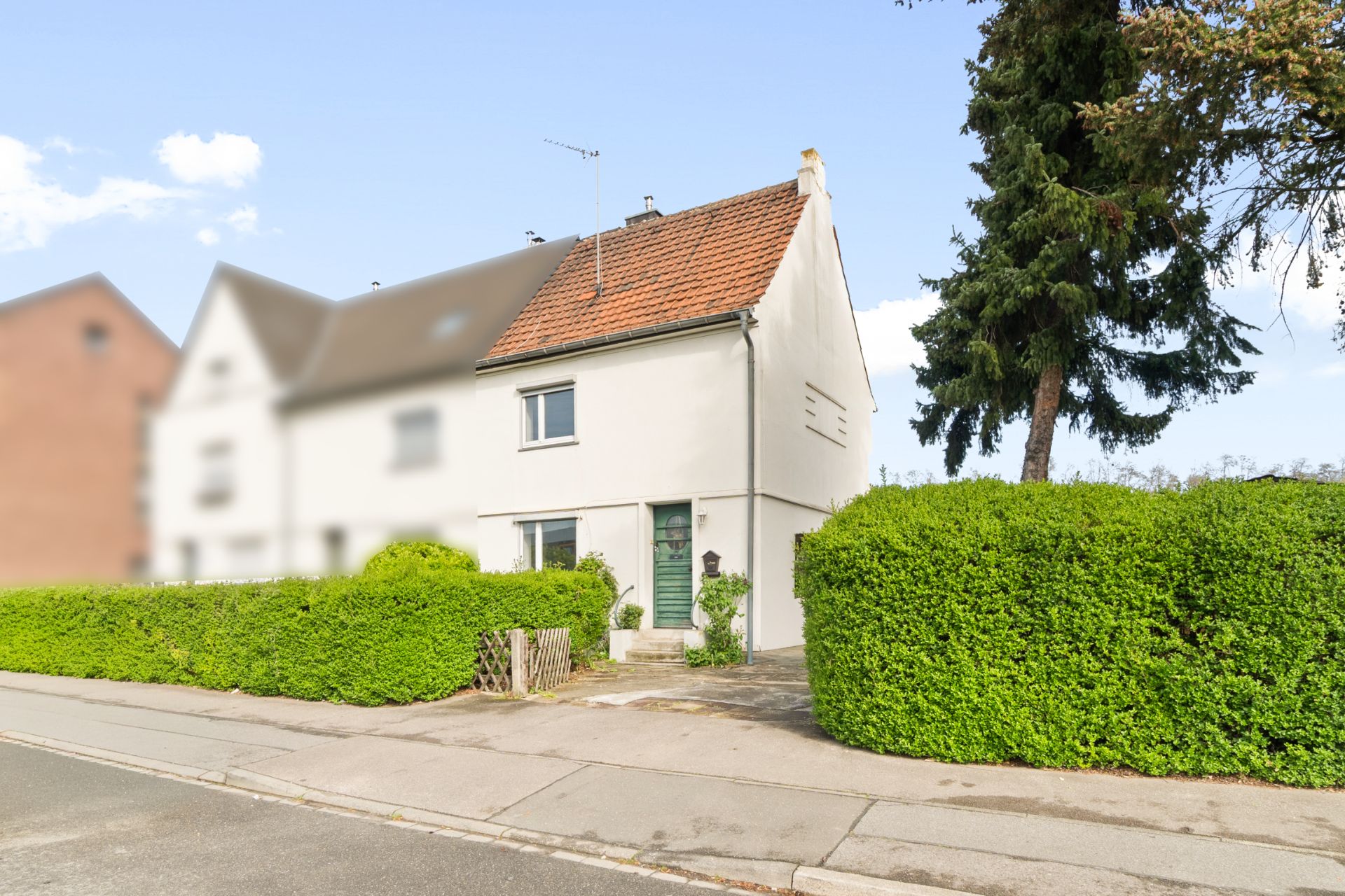 Weißes Einfamilienhaus mit rotem Dach, Garten und Hecke an einer ruhigen Straße.