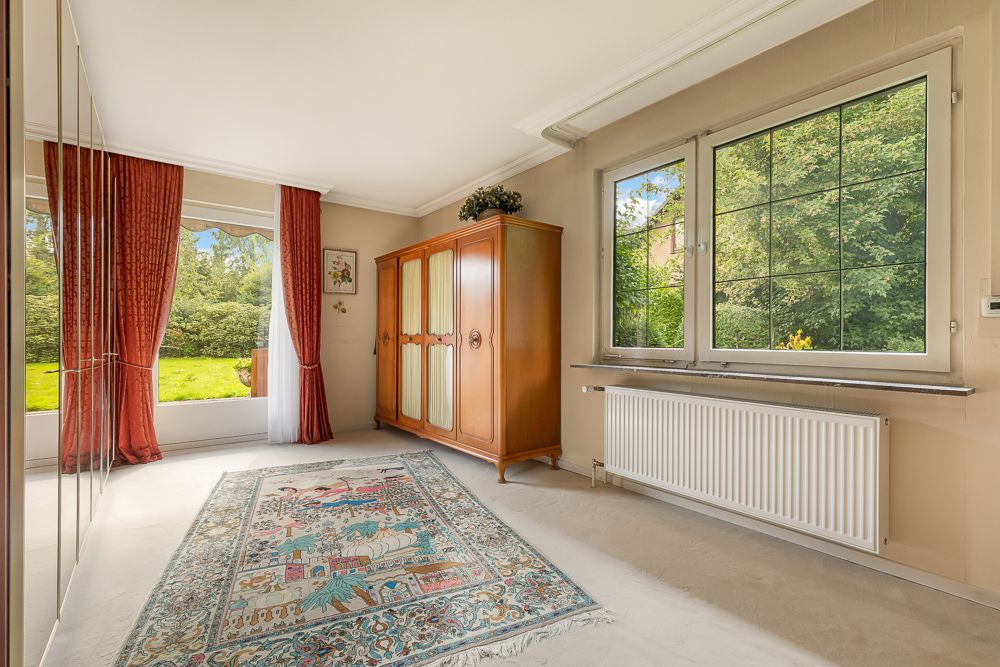 Helles Zimmer mit großem Fenster, rotem Vorhang, Teppich und Holzschrank, Blick ins Grüne.