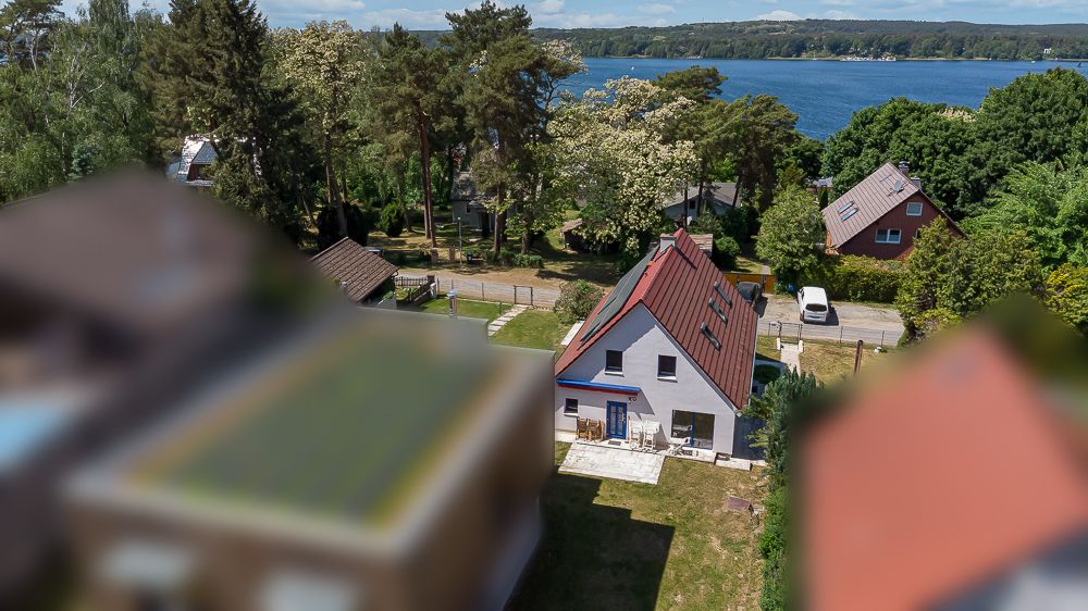 Haus mit rotem Dach und Garten nahe einem See, umgeben von Bäumen und weiteren Häusern.