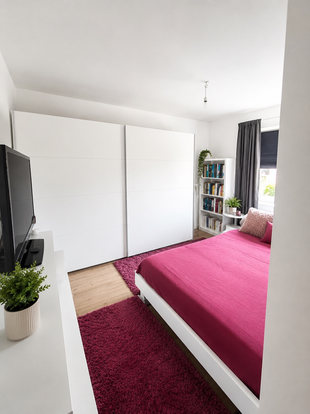 Modernes Schlafzimmer mit rotem Bett, Teppich, weißem Schrank, Fernseher und Bücherregal am Fenster.