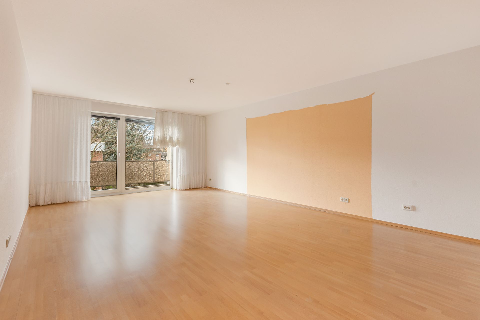 Leeres, helles Wohnzimmer mit Parkettboden, großer Fensterfront und Zugang zum Balkon.