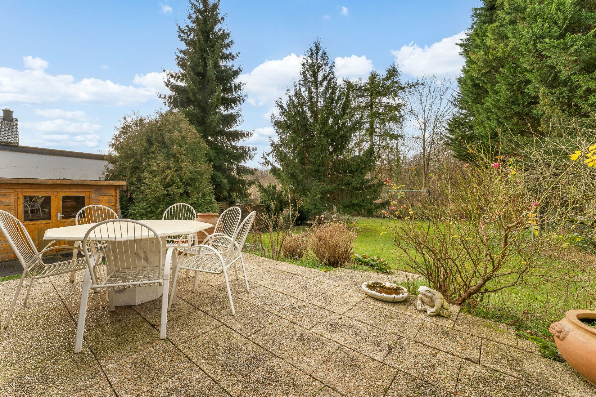 Steinterrasse mit rundem Tisch und Stühlen, Blick auf Garten und große Tannenbäume.