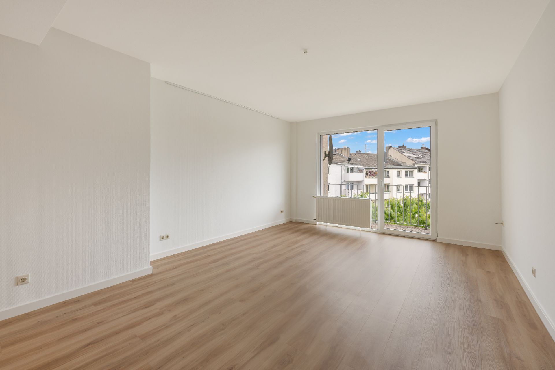 Leerer, heller Raum mit großem Fenster und Holzboden, Blick auf benachbarte Häuser.